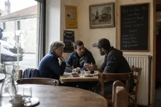 Three people of different generations conversing quietly at a table in a French village café with natural daylight, showing an authentic moment of intergenerational engagement.