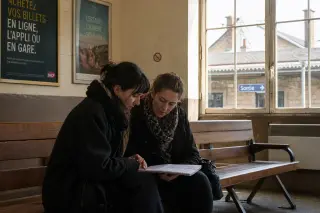 Two women in a French suburban train station waiting room, quietly discussing over a notebook in natural, soft daylight.