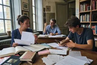 Three people—two adults and a teen—engaged in study and discussion at desks in a modest French library, with papers and books around, sunlight streaming softly through old windows.