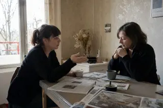 Two women in a lived-in French kitchen share a quiet, reflective morning conversation over coffee with natural light and subtle expression.