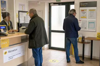 Two adults in a modest French post office interacting quietly, one at the counter, another reading a brochure, with soft daylight and a worn, everyday atmosphere.