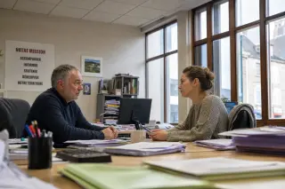 Two coworkers in a slightly cluttered rural French office quietly discussing work, natural morning light streaming through old windows, candid and authentic atmosphere.