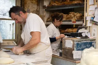Two bakery workers in a small, cluttered French bakery, one kneading dough and the other checking receipts, focused on their tasks in natural morning light.