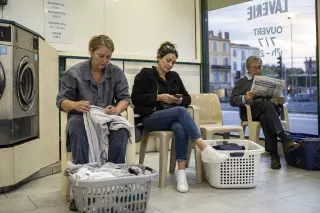 Three people in a French laundromat at dusk, two women sitting with laundry baskets and a man reading a newspaper, in a softly lit, realistic everyday scene.