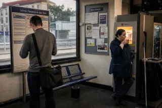 Two ordinary people in a modest French train station, one checking schedules, another sipping coffee near a vending machine, in gentle morning light.